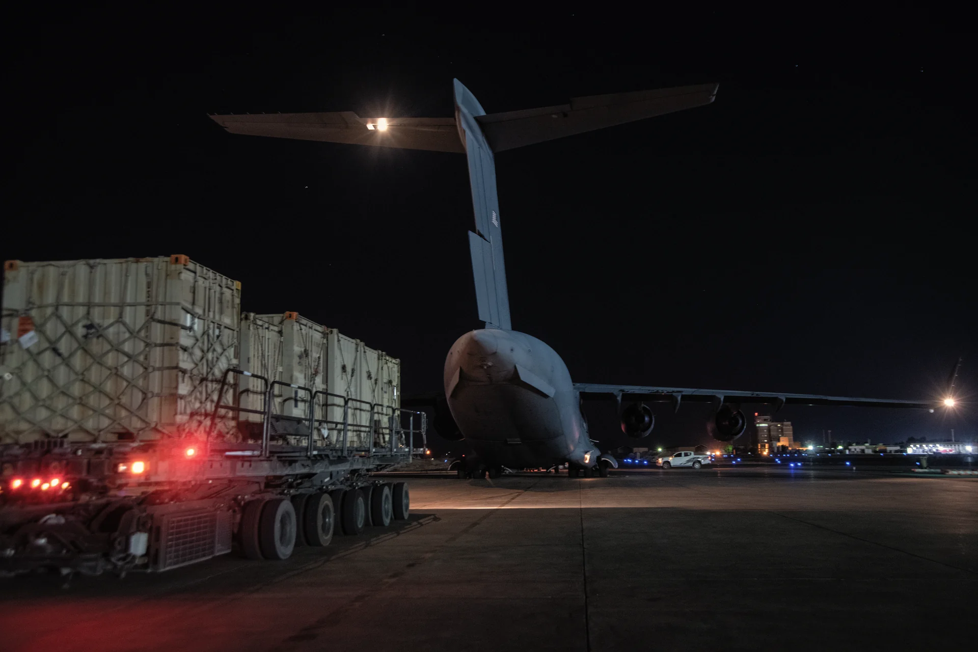 Netted 463L air cargo pallets on a cargo loader beside a military transport aircraft on the flight line at night